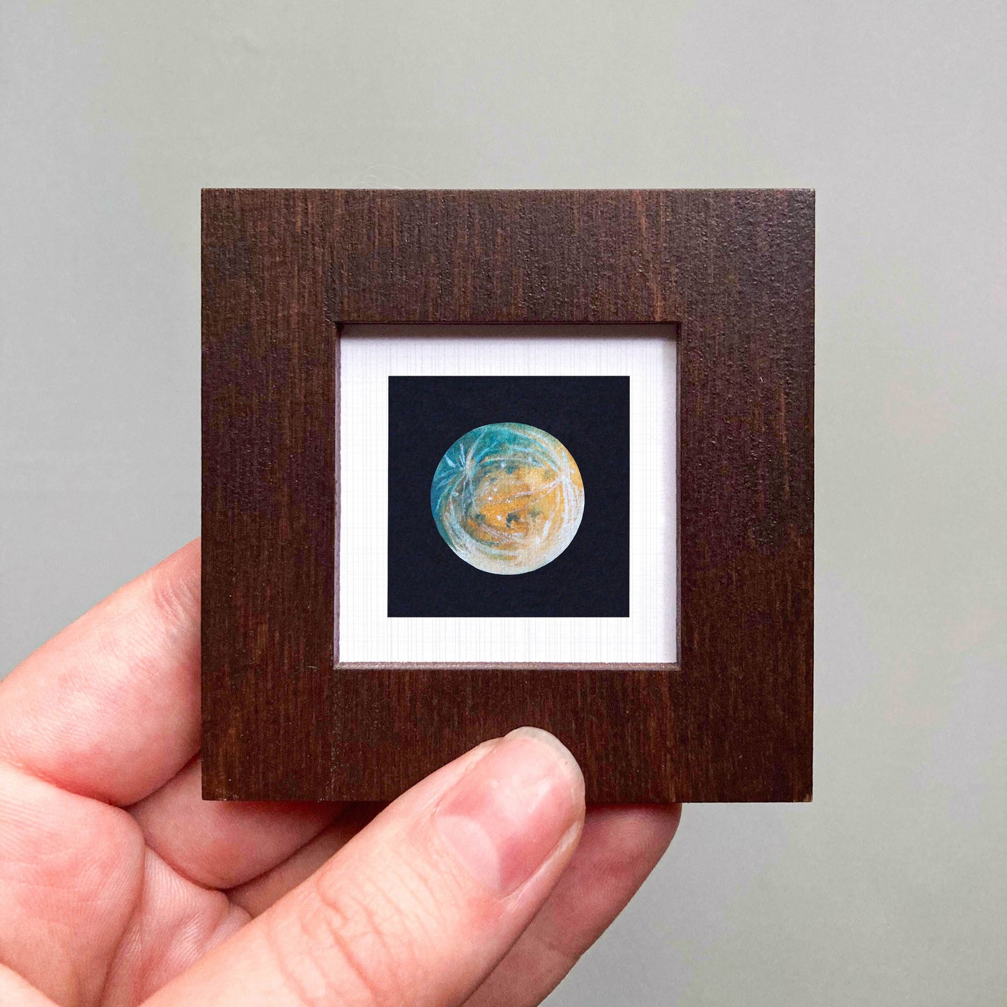 A hand is holding a small wooden frame containing a miniature painting of a planet.
