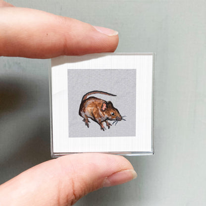 A person's hand is holding a small square picture of a brown mouse against a gray background.