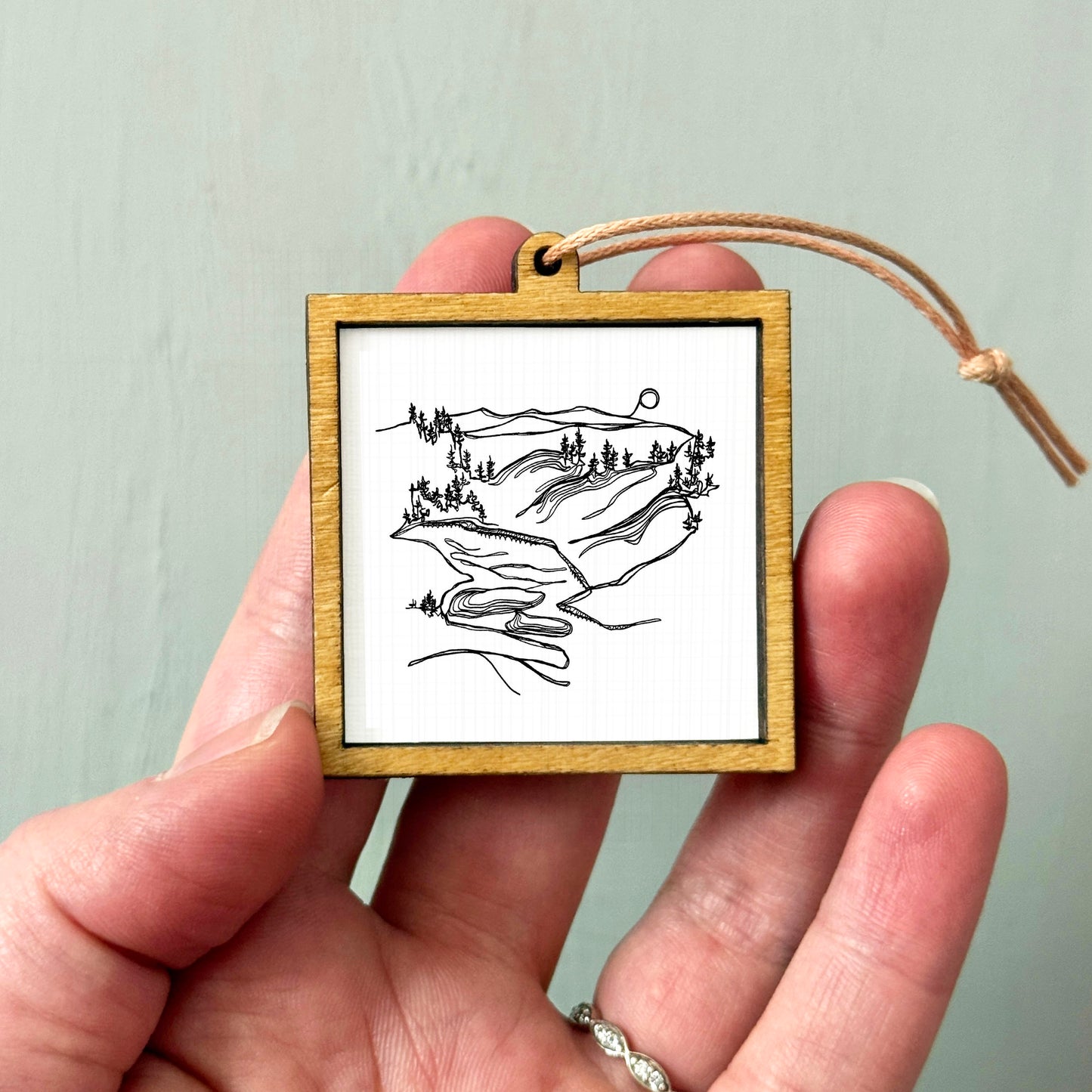 A hand holding a small wooden frame containing a black and white line drawing of a landscape with mountains and trees.