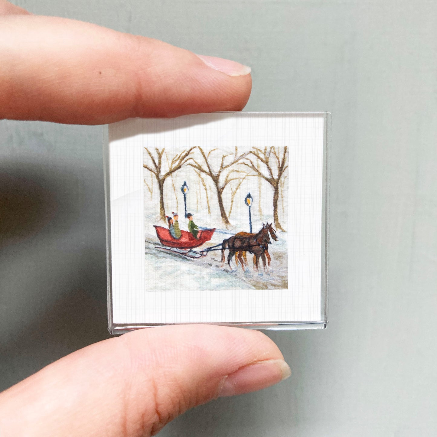 A person's hand is holding a small square picture depicting a red sleigh being pulled by a horse through a snowy landscape with bare trees.