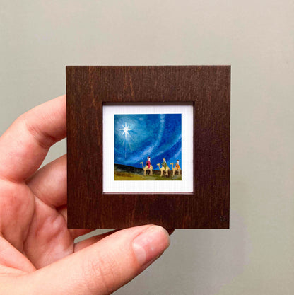 A hand is holding a small, square-shaped picture frame containing a painting of a starry night sky with a bright star and a group of people on a grassy field.