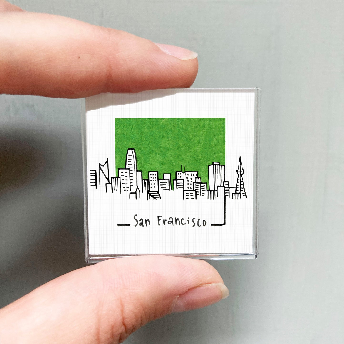 A hand holding a small square magnet with a green cityscape illustration on it.