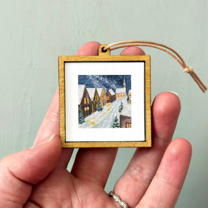 A hand holding a small wooden picture frame containing a winter village scene with snow-covered houses and a church.