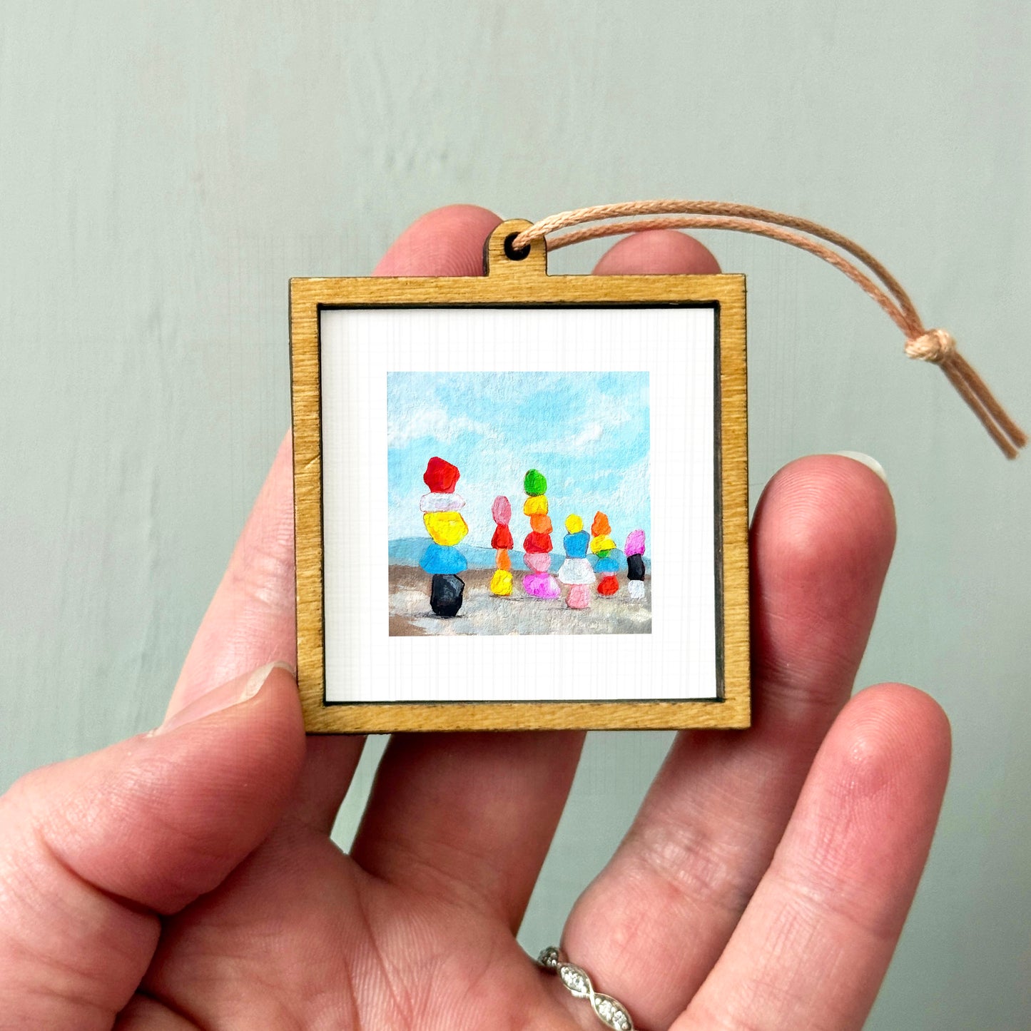 A hand holding a small wooden frame containing a painting of colorful stacked objects against a blue sky background.