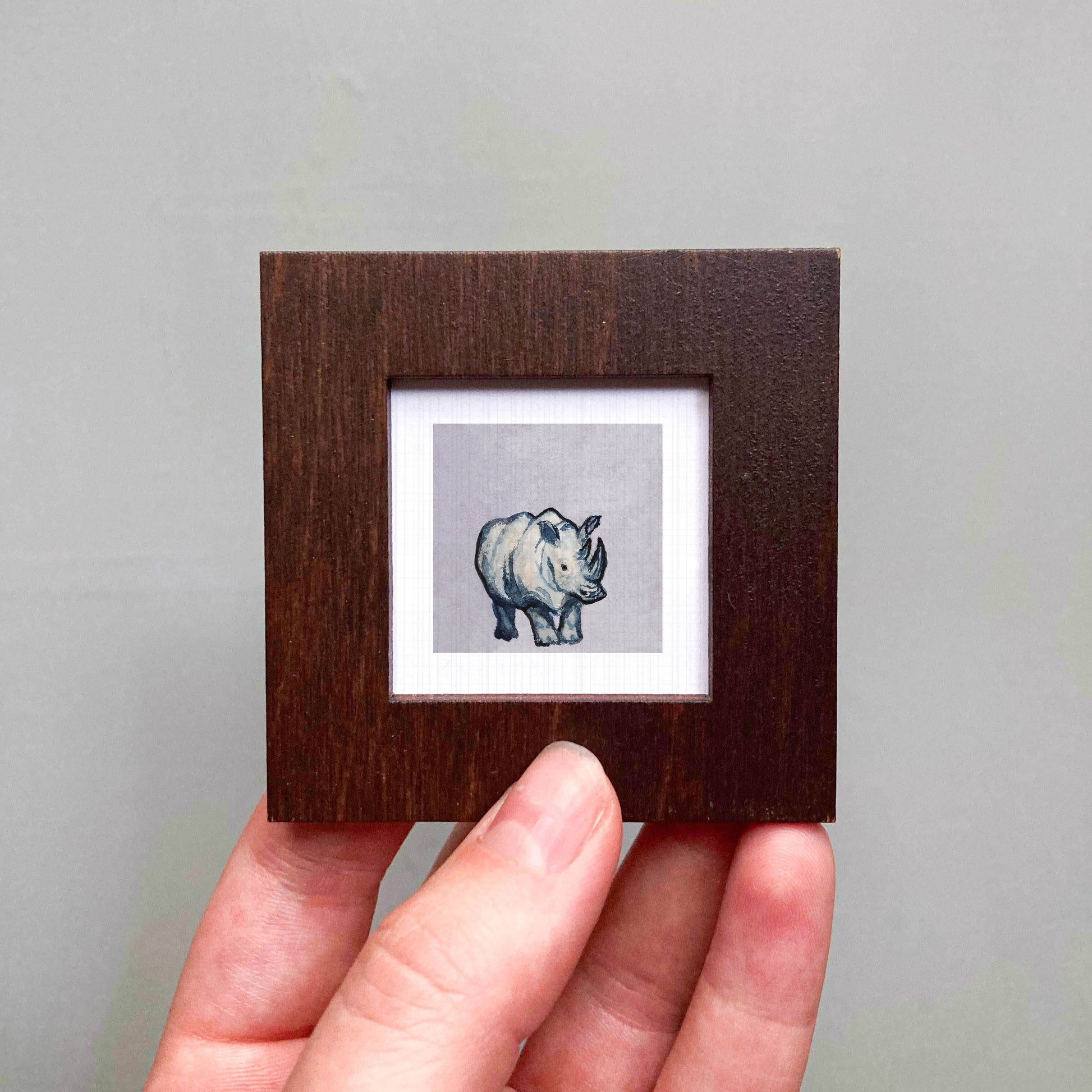 A person's hand is holding a small wooden frame containing a single, detailed illustration of a rhinoceros.