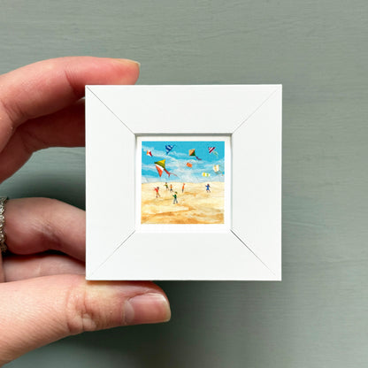 A hand is holding a small white picture frame containing a painting of people flying kites on a beach.