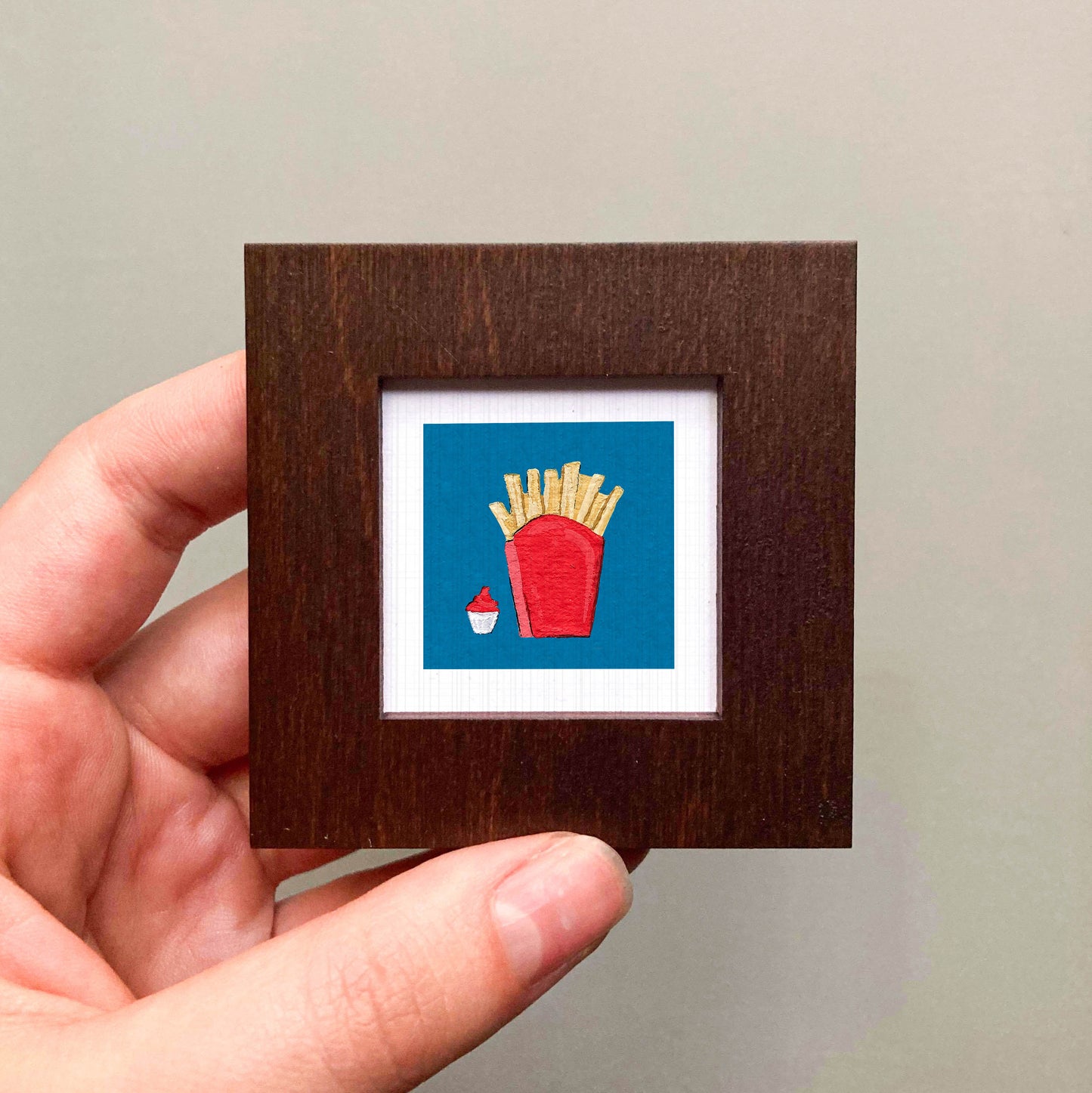 A hand is holding a small wooden frame containing a framed image of a red cup with french fries inside.