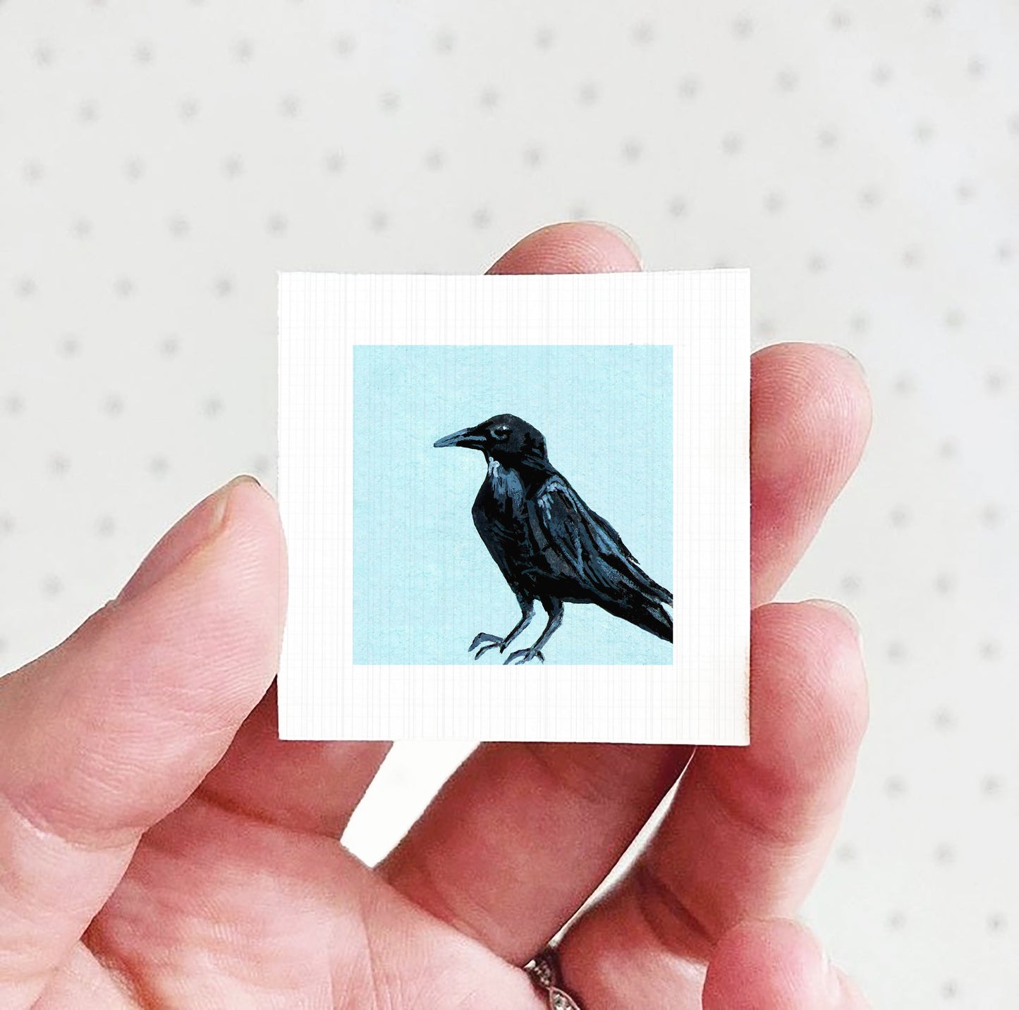 A hand holding a small square picture of a black crow against a light blue background.