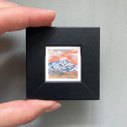A person's hand holding a small black picture frame containing a miniature painting of a mountain landscape with a colorful sky.