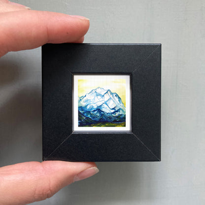 A person's hand is holding a small black frame containing a framed painting of a mountain landscape.