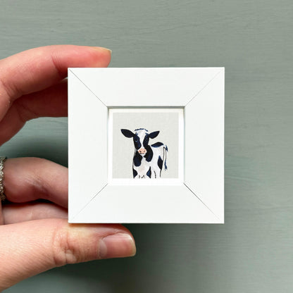 A hand is holding a small white picture frame containing a black and white cow image.