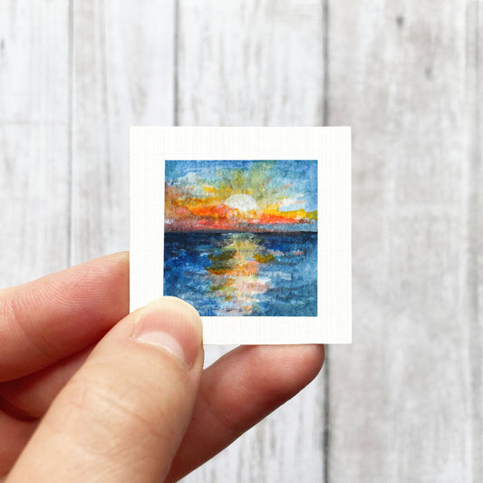 A hand is holding a small square painting of a sunset over a body of water.