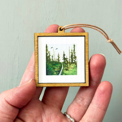A hand holding a small wooden frame containing a painting of a forest scene.
