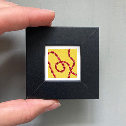 A person's hand is holding a small black square frame containing a vibrant yellow and red abstract artwork.