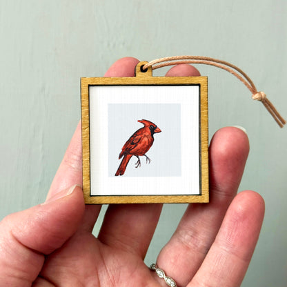 A person's hand holding a small wooden frame containing a framed image of a red cardinal bird.