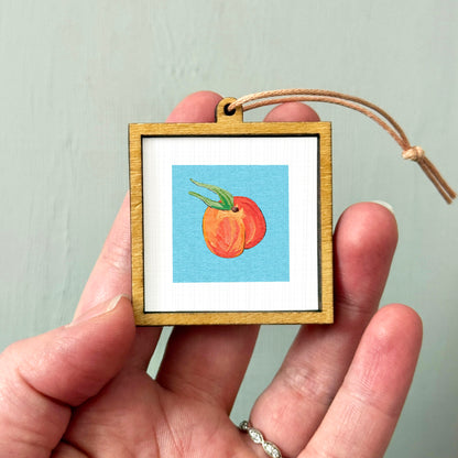 A person's hand holding a small wooden frame containing a framed image of a peach.