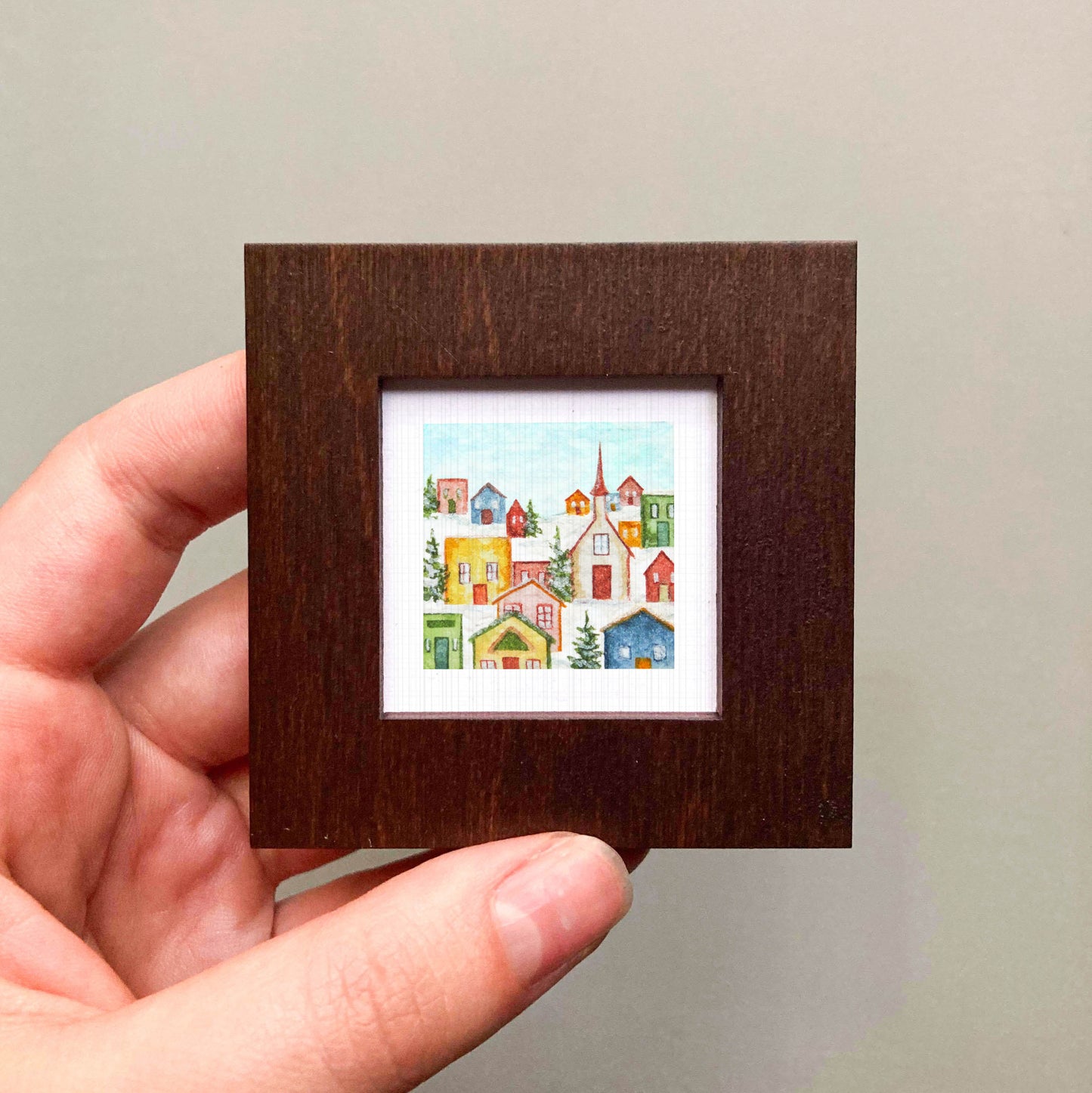 A hand is holding a small, square-shaped picture frame containing a colorful, miniature painting of a village scene.