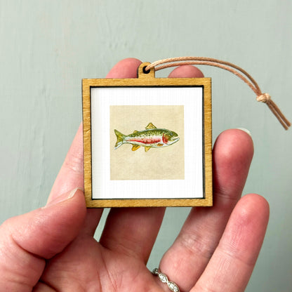 A person's hand holding a small wooden frame containing a framed illustration of a fish.