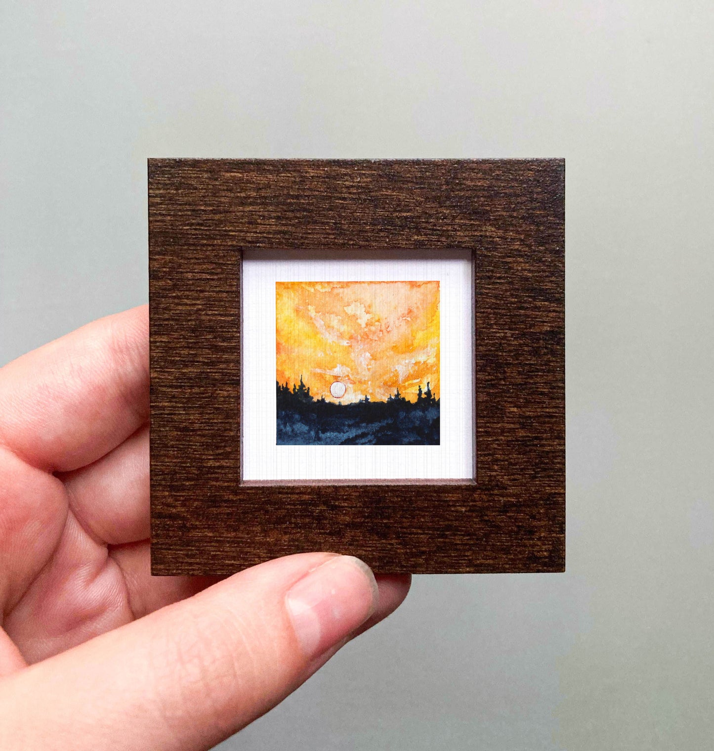 A hand is holding a small, square-shaped wooden frame containing a miniature painting of a sunset landscape.