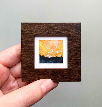 A hand is holding a small, square-shaped wooden frame containing a miniature painting of a sunset landscape.