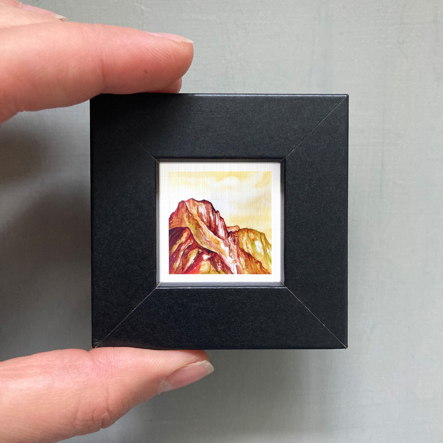 A person's hand holding a small black frame containing a framed painting of a mountain landscape.