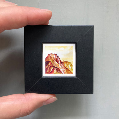 A person's hand holding a small black frame containing a framed painting of a mountain landscape.