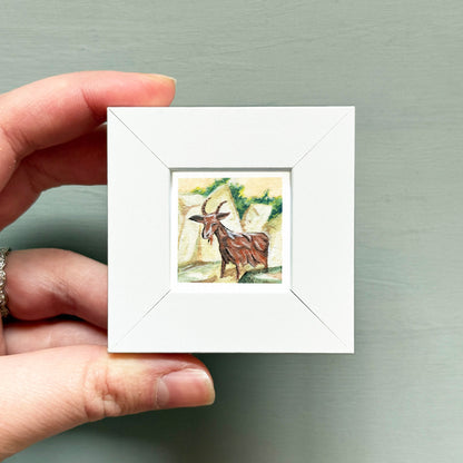 A hand is holding a small white square frame containing a painting of a brown goat.