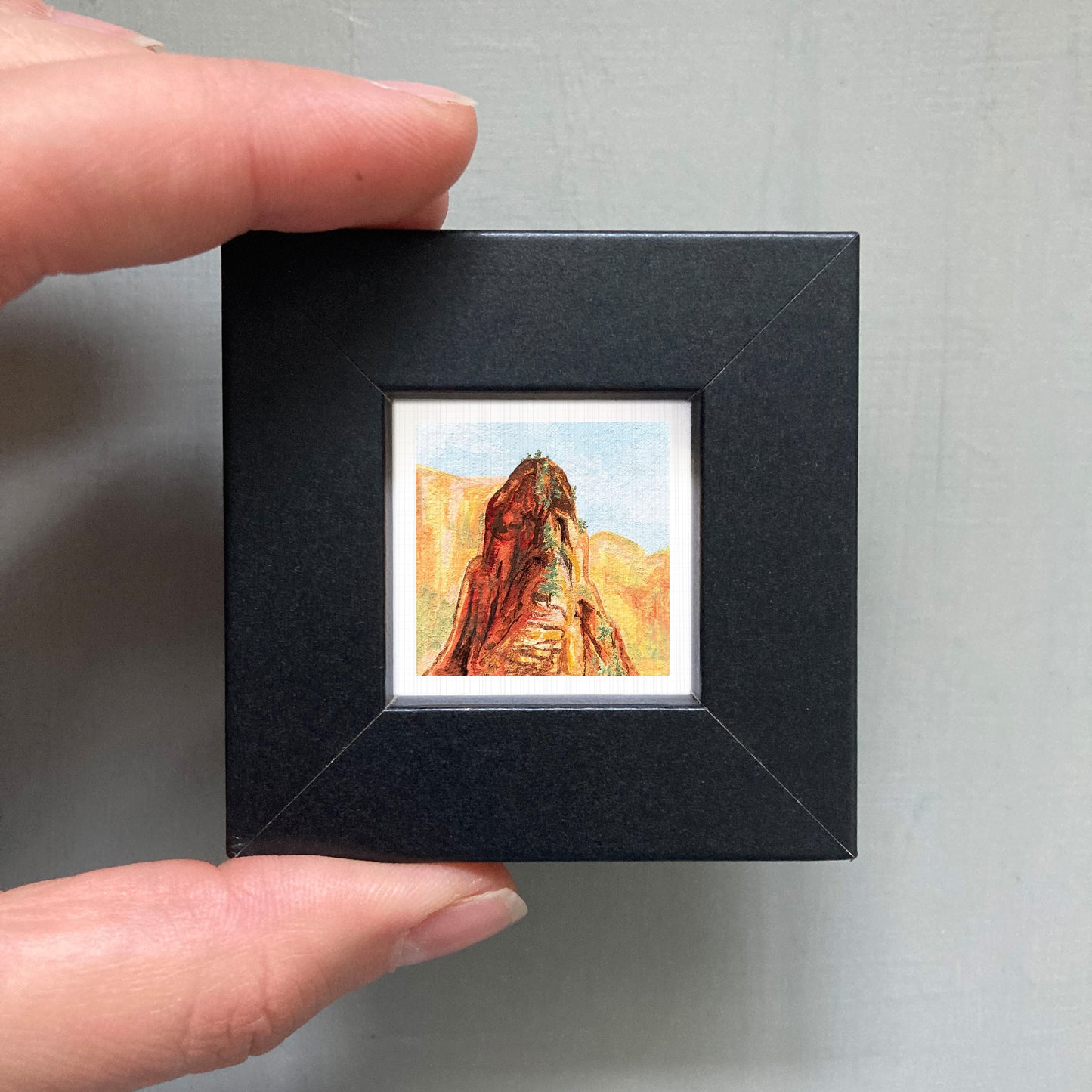 A person's hand holding a small black frame containing a framed painting of a mountain landscape.