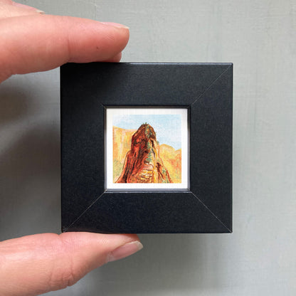 A person's hand holding a small black frame containing a framed painting of a mountain landscape.