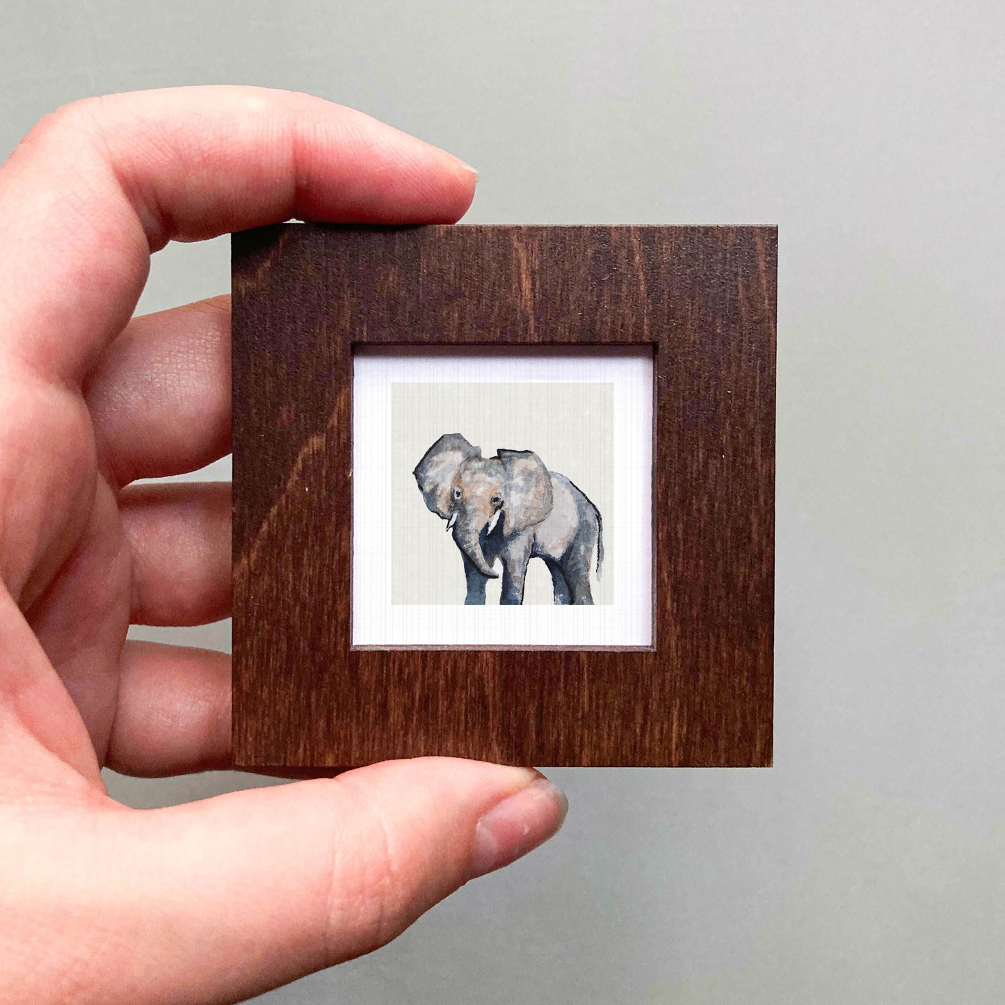 A hand is holding a small wooden frame containing a picture of an elephant.