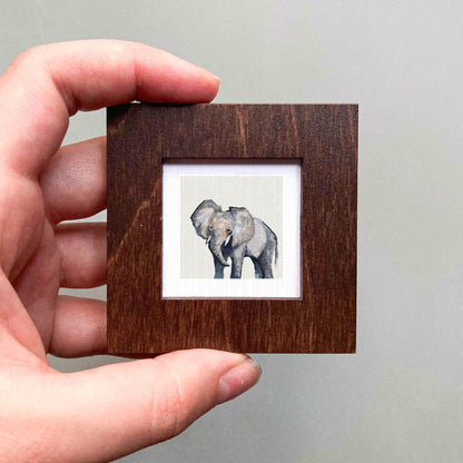 A hand is holding a small wooden frame containing a picture of an elephant.