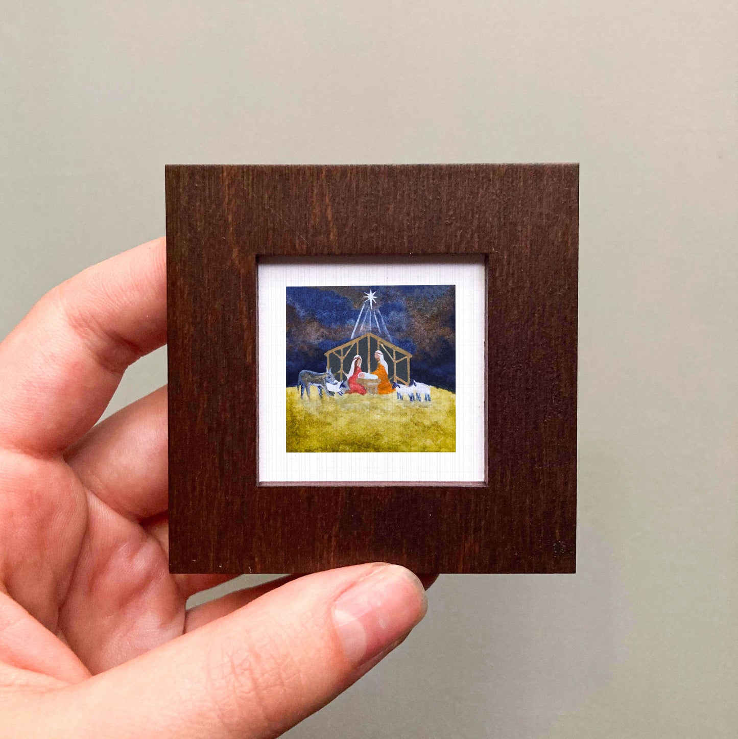 A hand is holding a small, square-shaped picture frame containing a painting of a nativity scene.