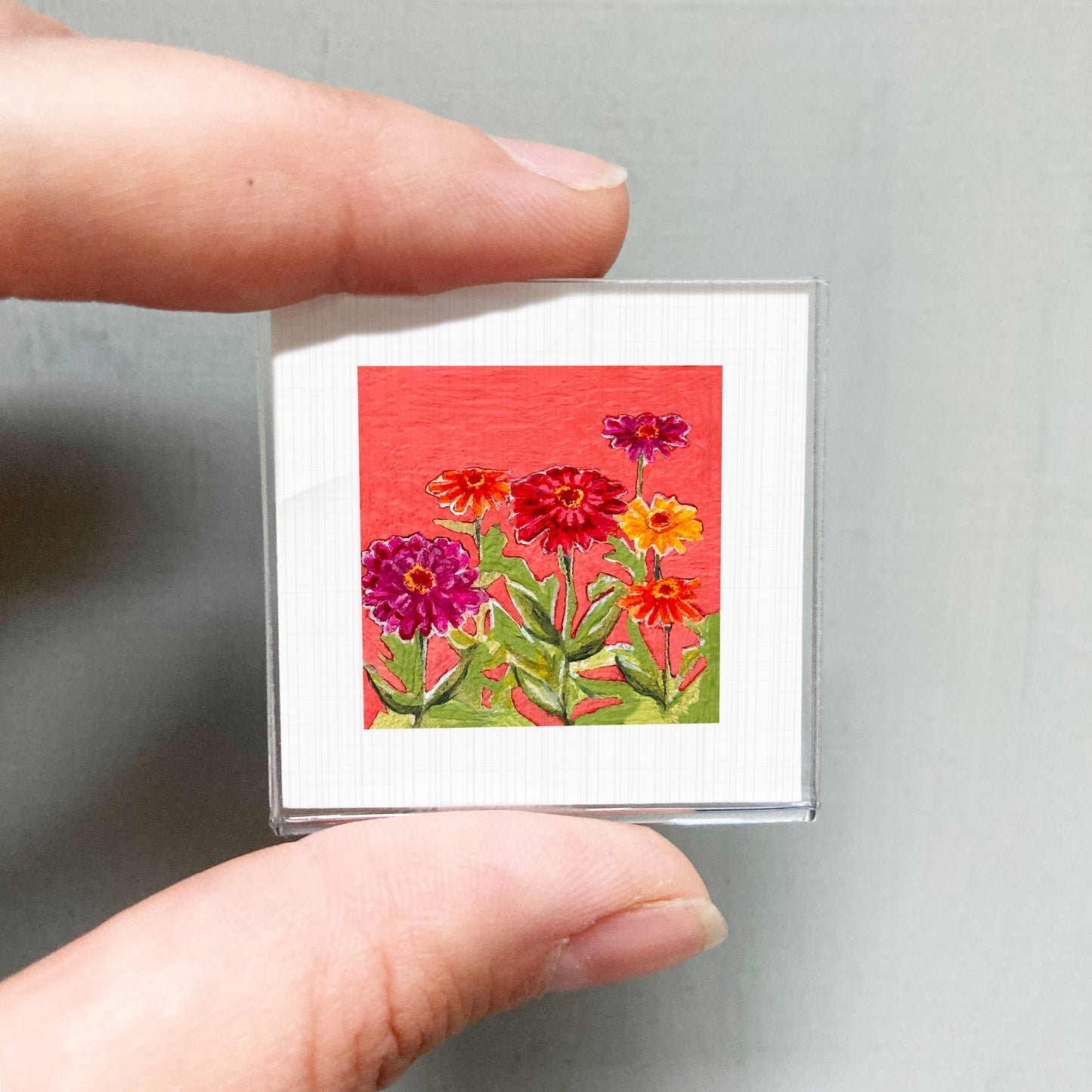 A person's hand is holding a small square card with a vibrant painting of colorful flowers against a red background.