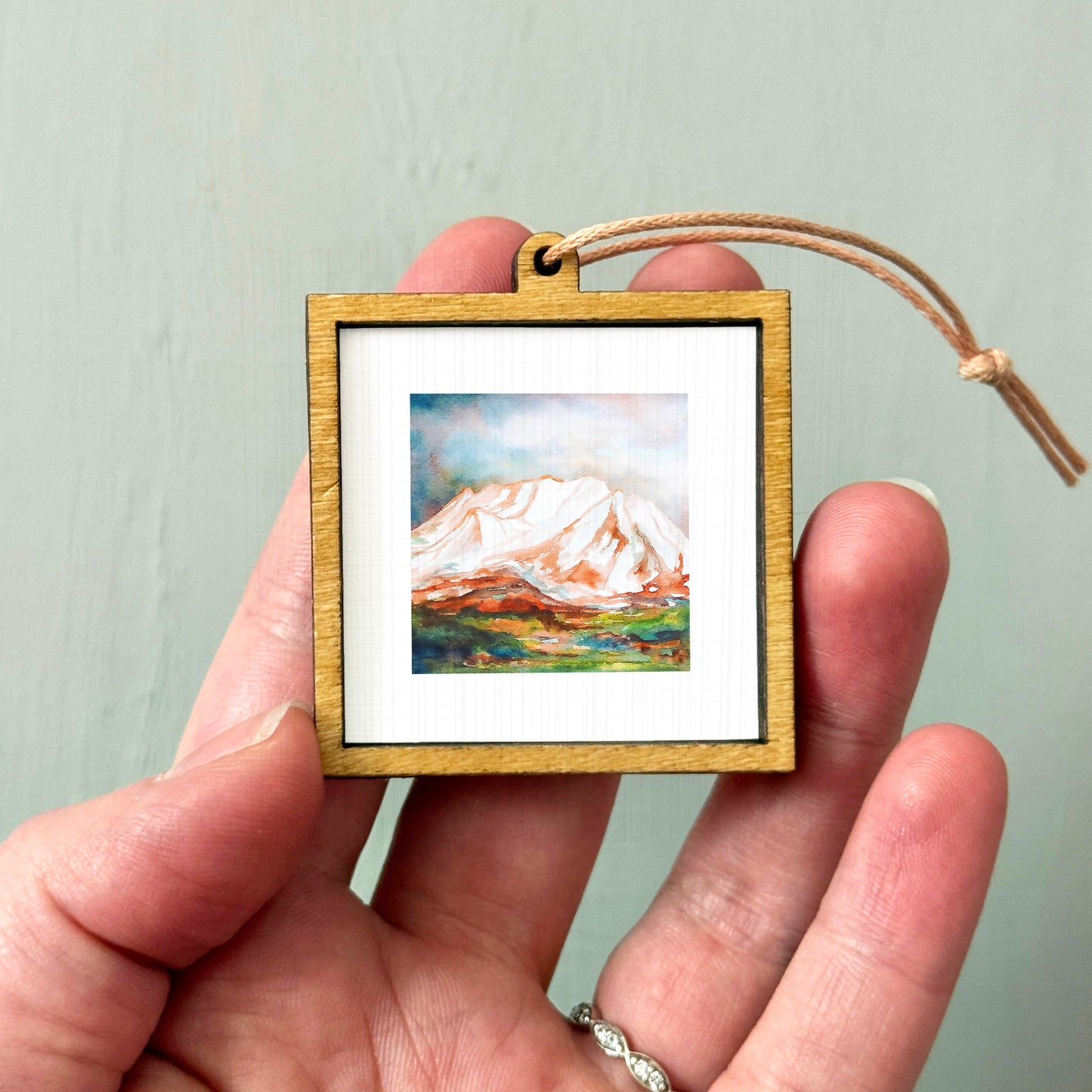 A hand holding a small wooden frame containing a painting of a mountain landscape.