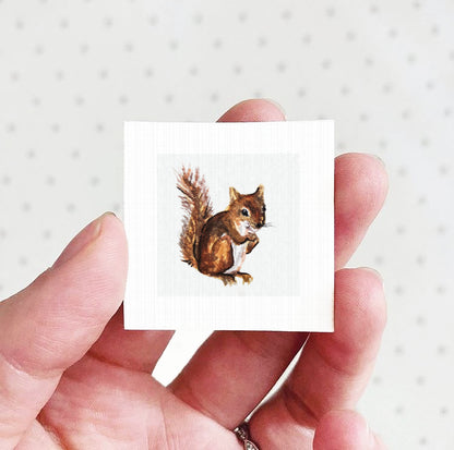 A hand holding a small square card with a painting of a squirrel on it.
