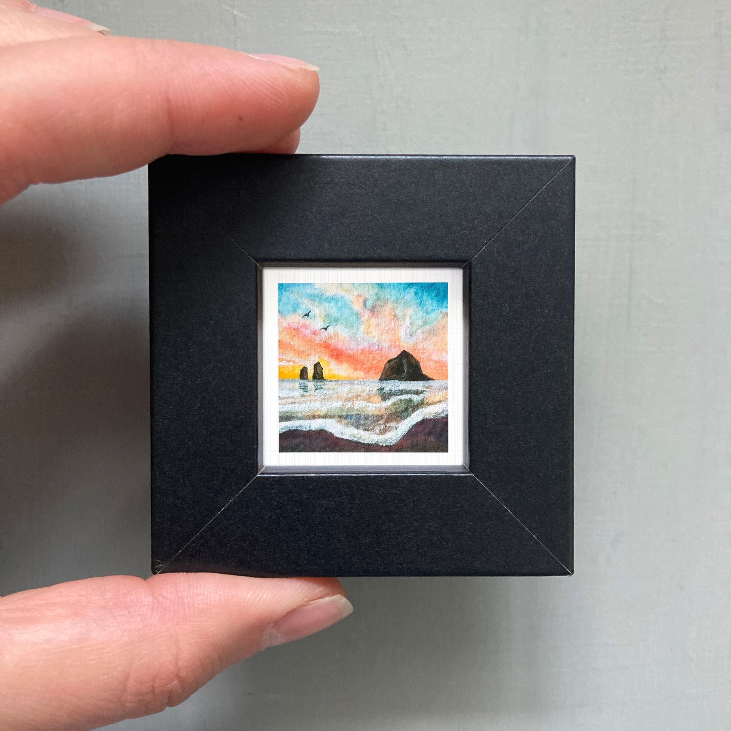 A person's hand holding a small, square-shaped black frame containing a colorful painting of a beach scene with a rock formation in the background.