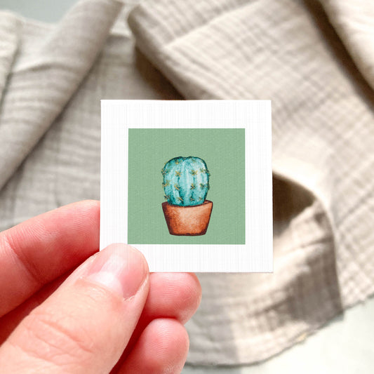 A hand is holding a small square card with a picture of a cactus on it.