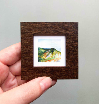 A hand is holding a small, square-shaped wooden frame containing a watercolor painting of a mountain landscape.