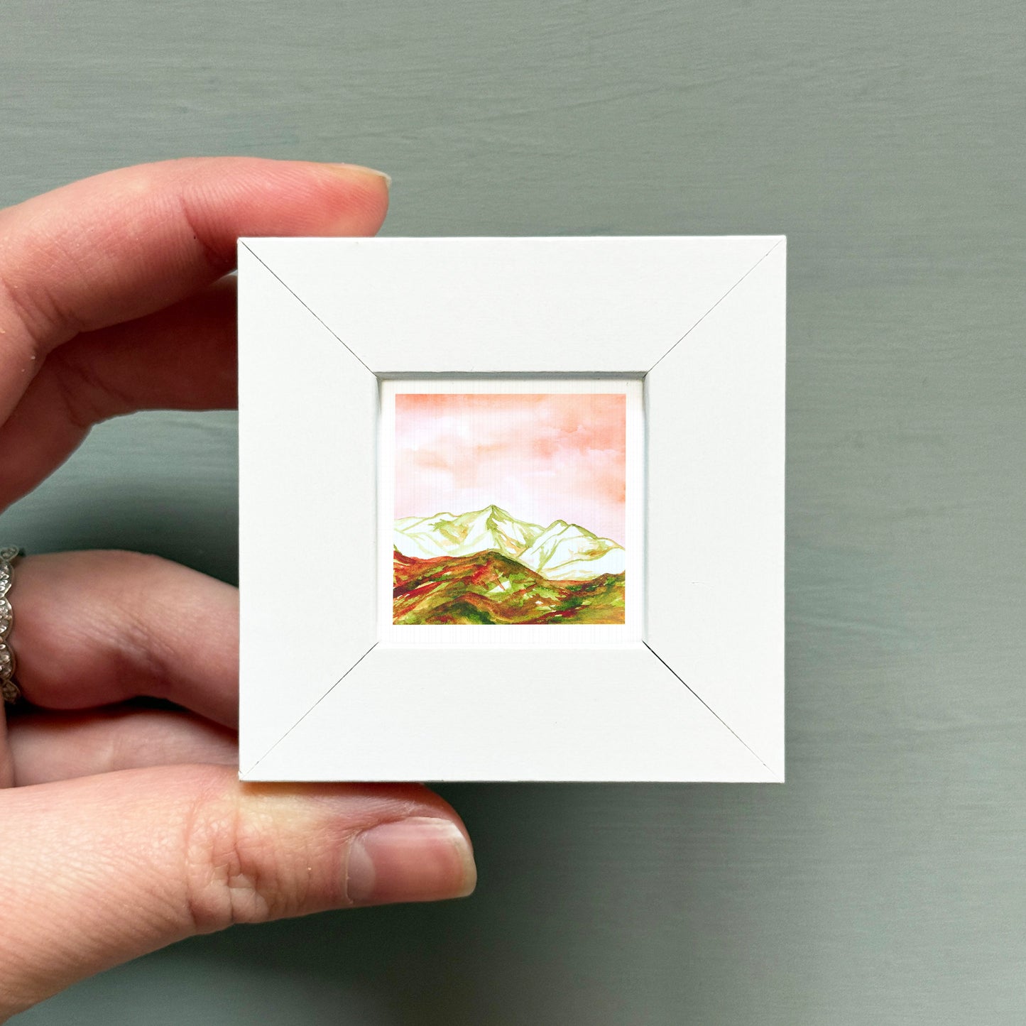 A hand is holding a small white picture frame containing a landscape painting of mountains.