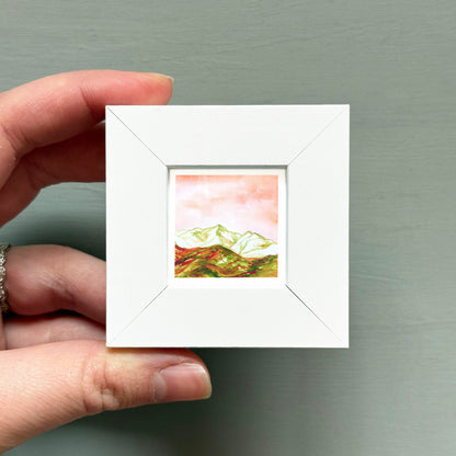 A hand is holding a small white picture frame containing a landscape painting of mountains.