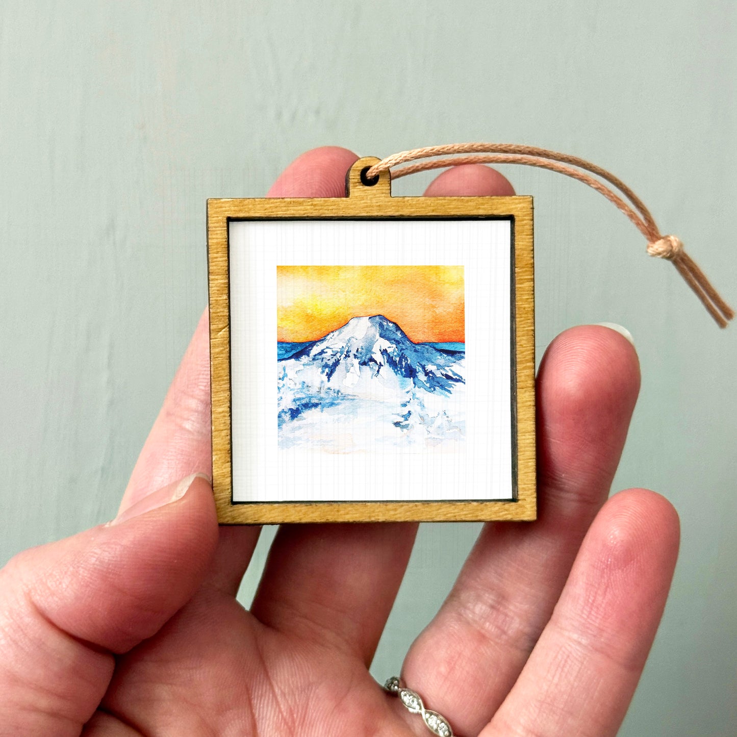 A hand holding a small wooden frame containing a watercolor painting of a mountain landscape.