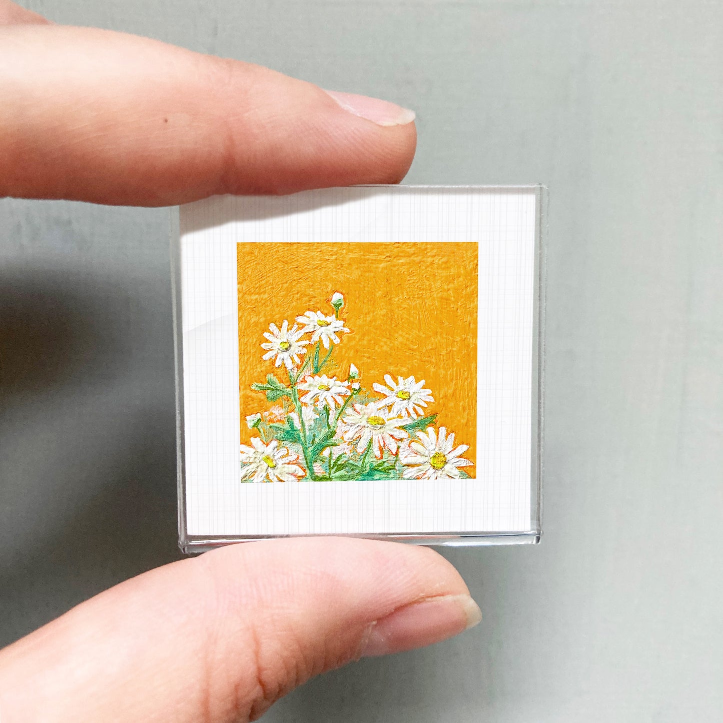 A person's hand is holding a small square picture of white daisies against an orange background.