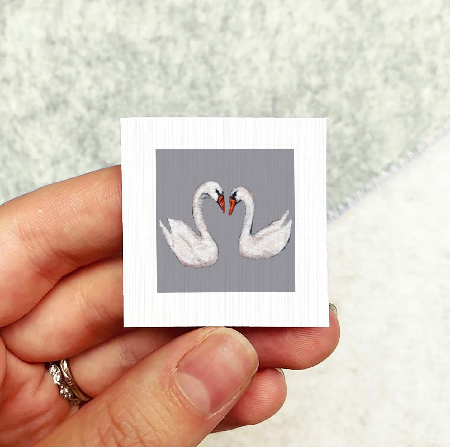 A hand holding a small square picture of two swans.