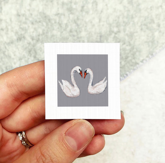 A hand holding a small square picture of two swans.