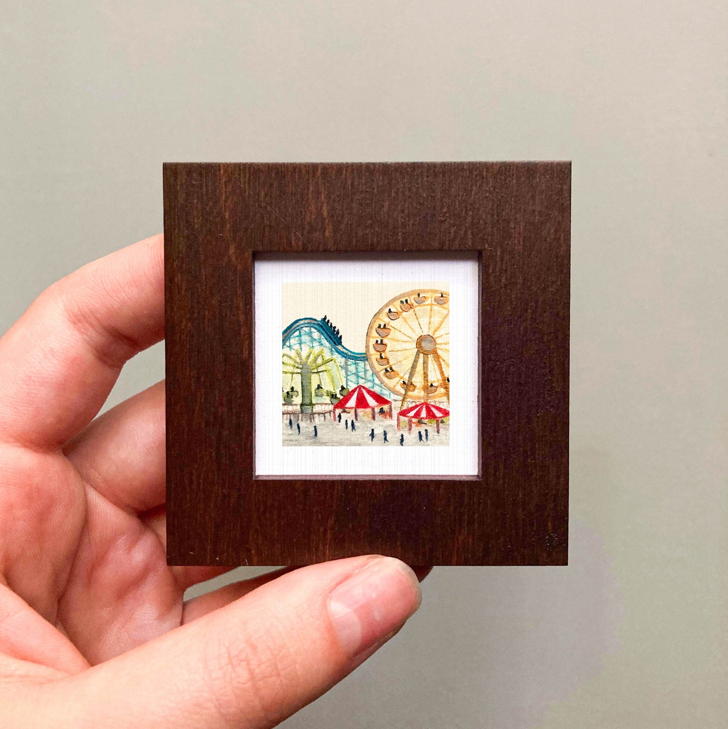 A hand is holding a small, square-shaped picture frame containing an illustration of a carnival scene with ferris wheels and other rides.