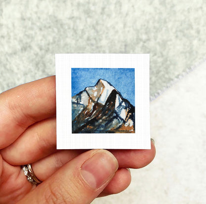 A person's hand holding a small square artwork depicting a mountain landscape.