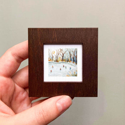 A hand is holding a small, square-shaped wooden frame containing a painting of people ice skating on a frozen lake.