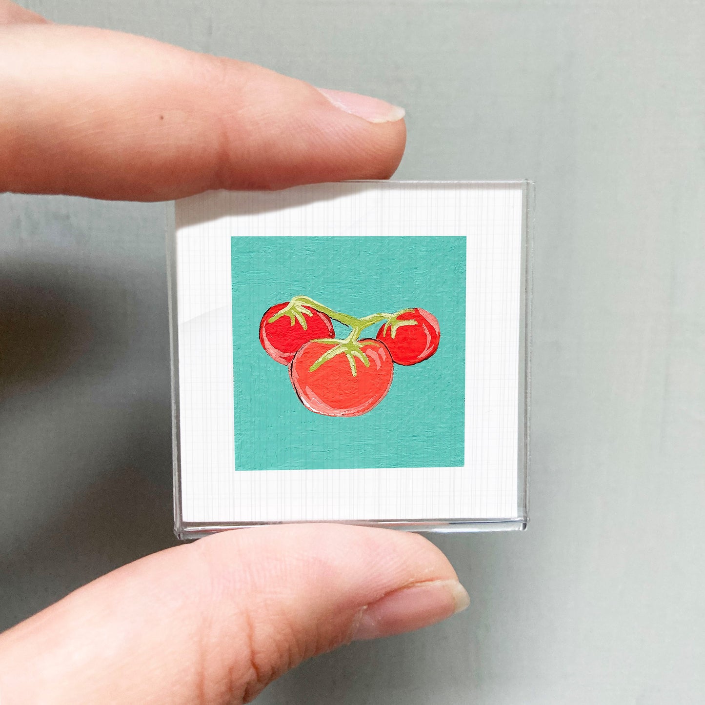 A person's hand holding a small square with a vibrant illustration of three red tomatoes on a green background.