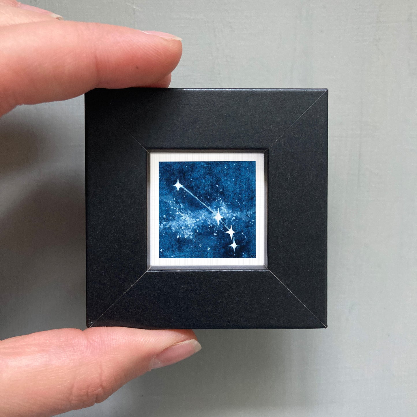 A hand is holding a small black frame containing a framed image of a starry night sky.