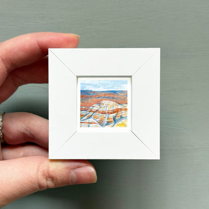 A hand is holding a small white square frame containing a painting of a landscape with mountains and a blue sky.